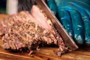Barbecue brisket being expertly sliced by a gloved hand on a wooden cutting board.
